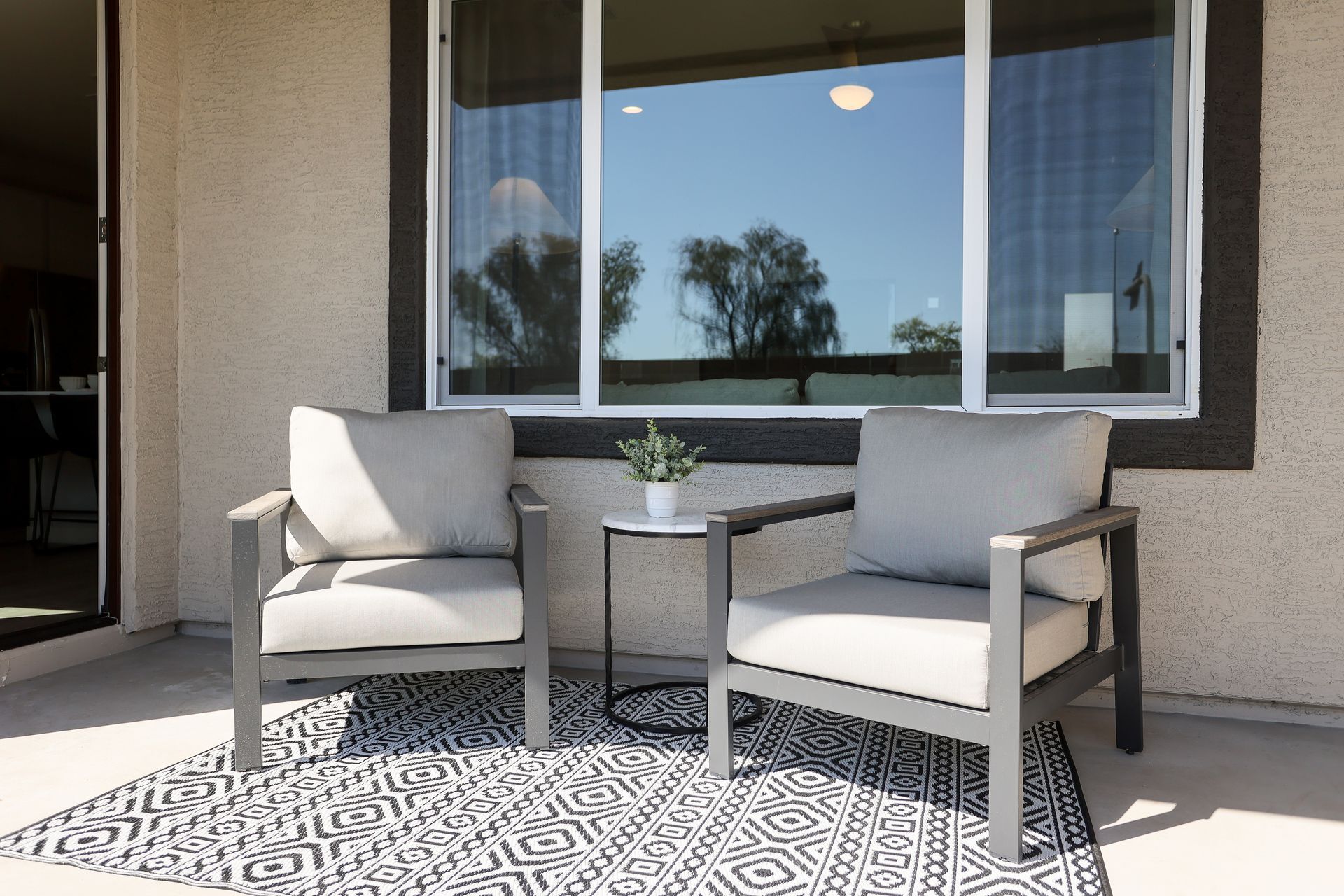 Two gray patio chairs flanking a small table on a patterned rug outside a window.