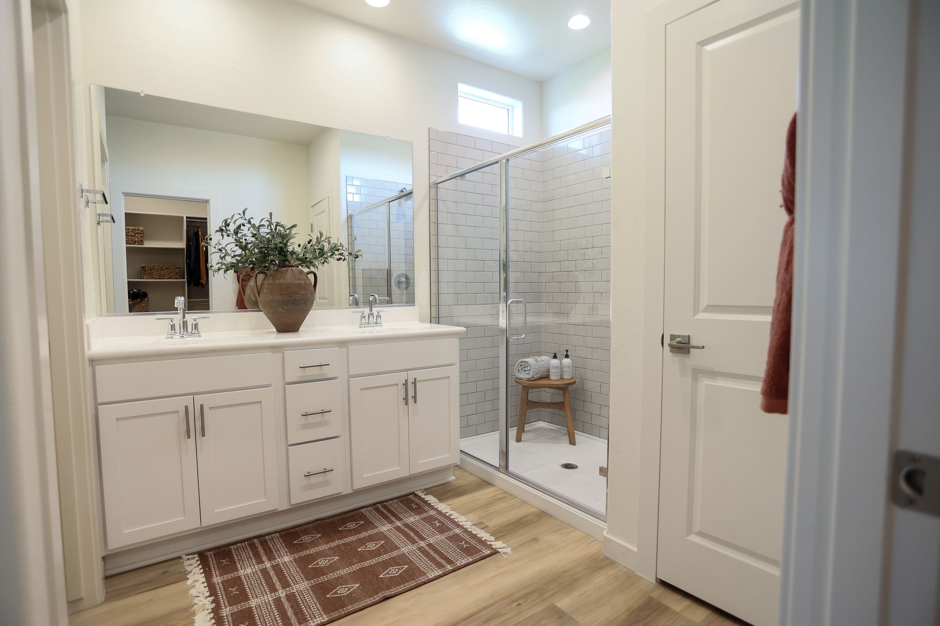 Bright bathroom with white vanity, glass shower, wood floor, and patterned rug