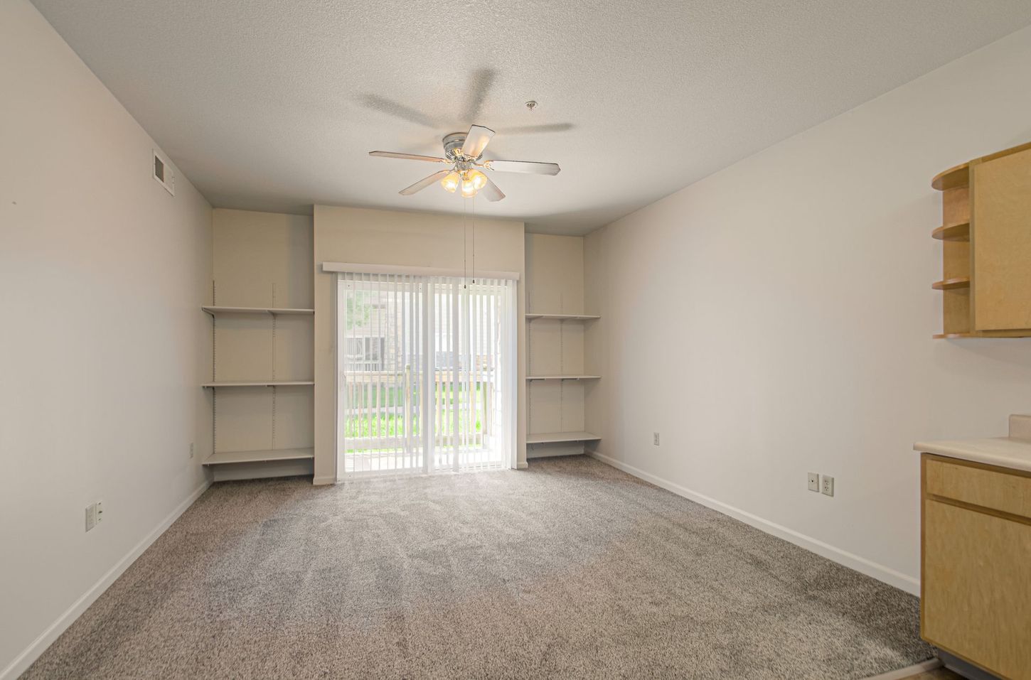 Empty room with carpet, sliding glass door to balcony, built-in shelves, and a ceiling fan.