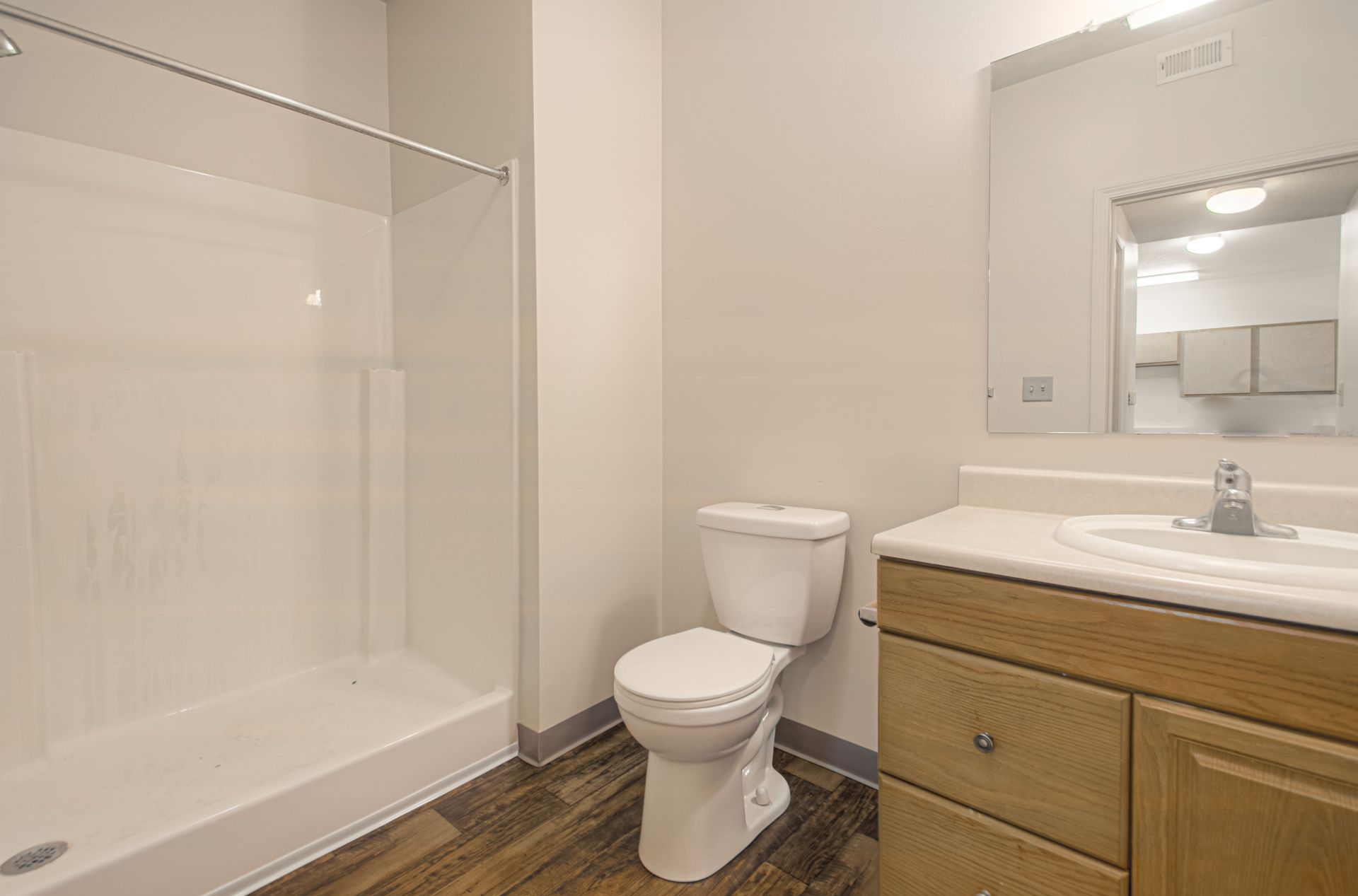 A modern bathroom with a walk-in shower, toilet, vanity, and large mirror; neutral tones with wood-look flooring.