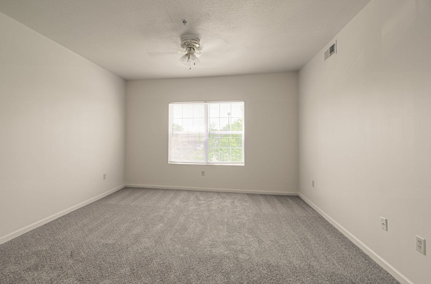 Empty bedroom with gray carpet, white walls, window, and ceiling fan.