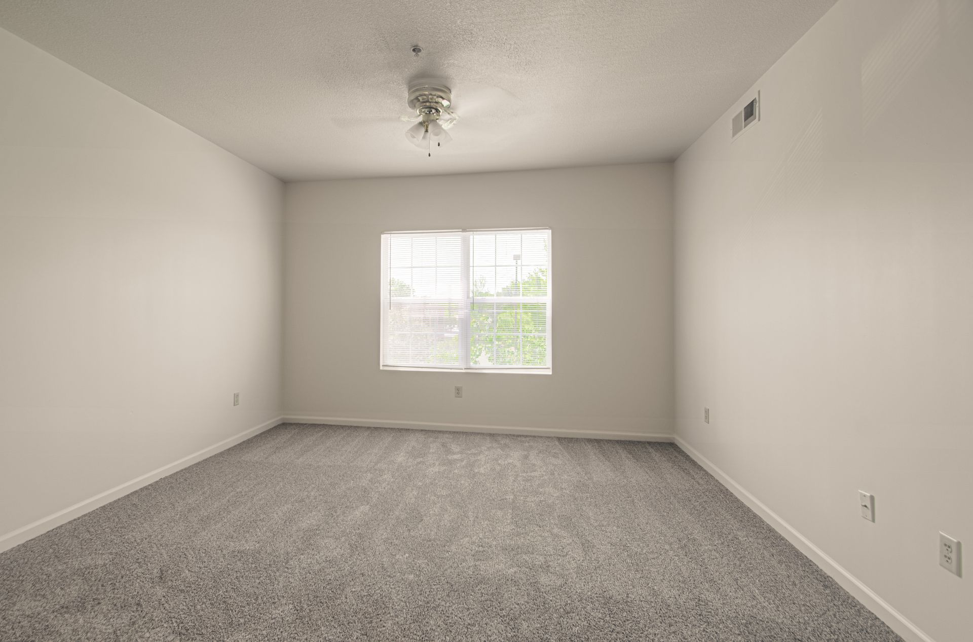 Empty bedroom with gray carpet, white walls, window, and ceiling fan.