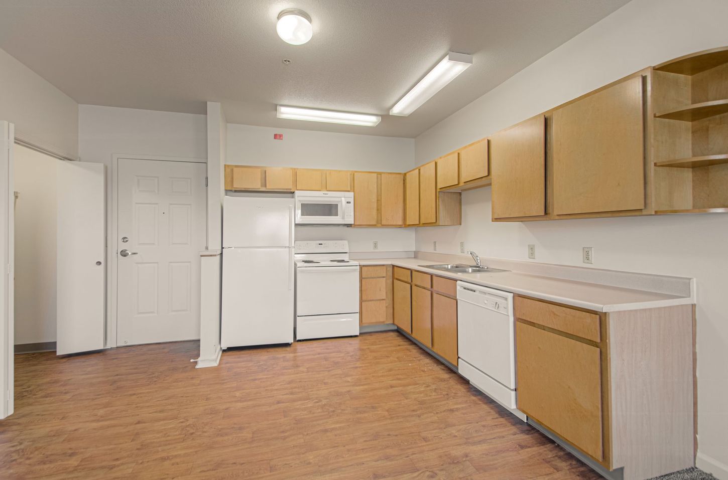 A small kitchen with light brown cabinets, white appliances, and tan flooring. A door is on the left.