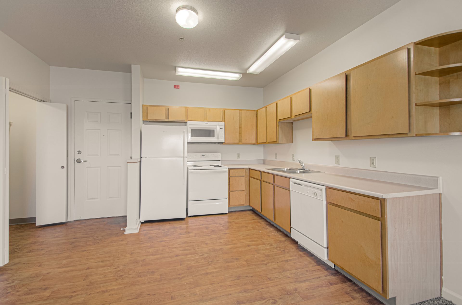 A small kitchen with light brown cabinets, white appliances, and tan flooring. A door is on the left.