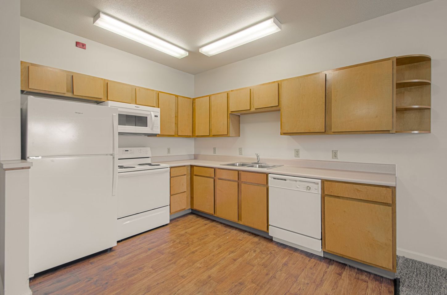 A kitchen with light wooden cabinets, white appliances, and a faux wood floor. It is brightly lit.