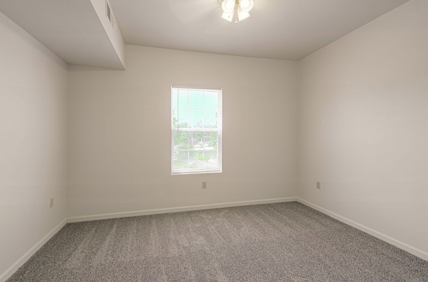 Empty bedroom with gray carpet, white walls, and a window. Ceiling light fixture and a small window.