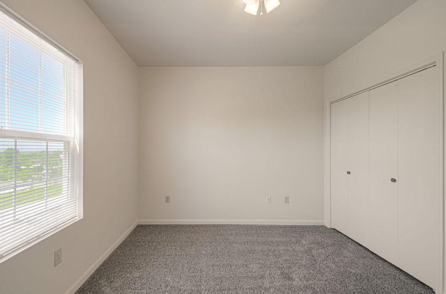Empty bedroom with gray carpet, white walls, a closed closet, and a window with blinds.