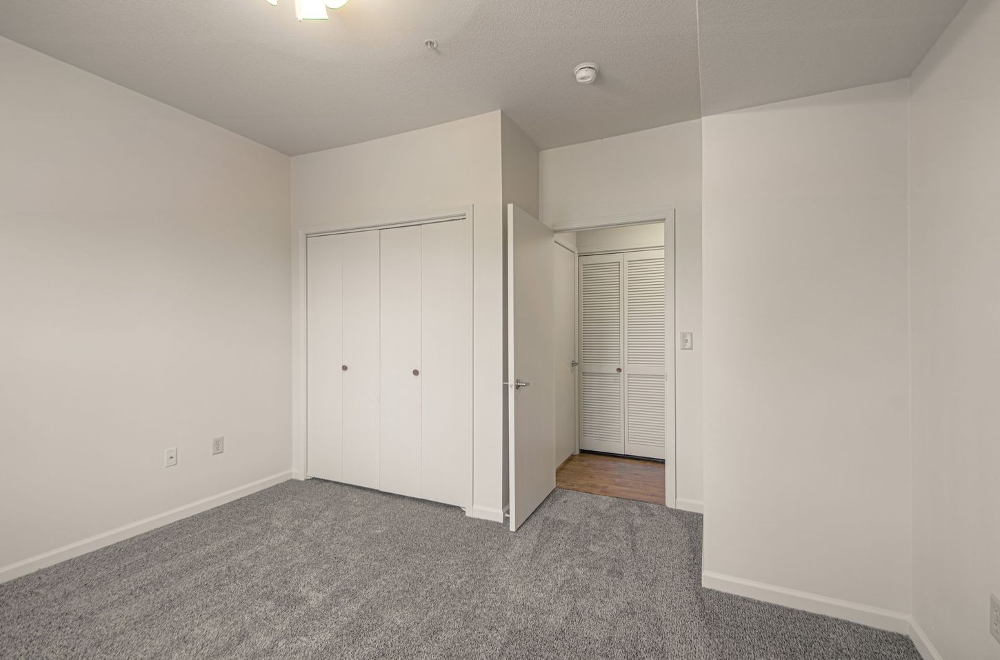 Empty bedroom with gray carpet, white walls, a closed closet, and an open door leading to a hallway with a folding door.