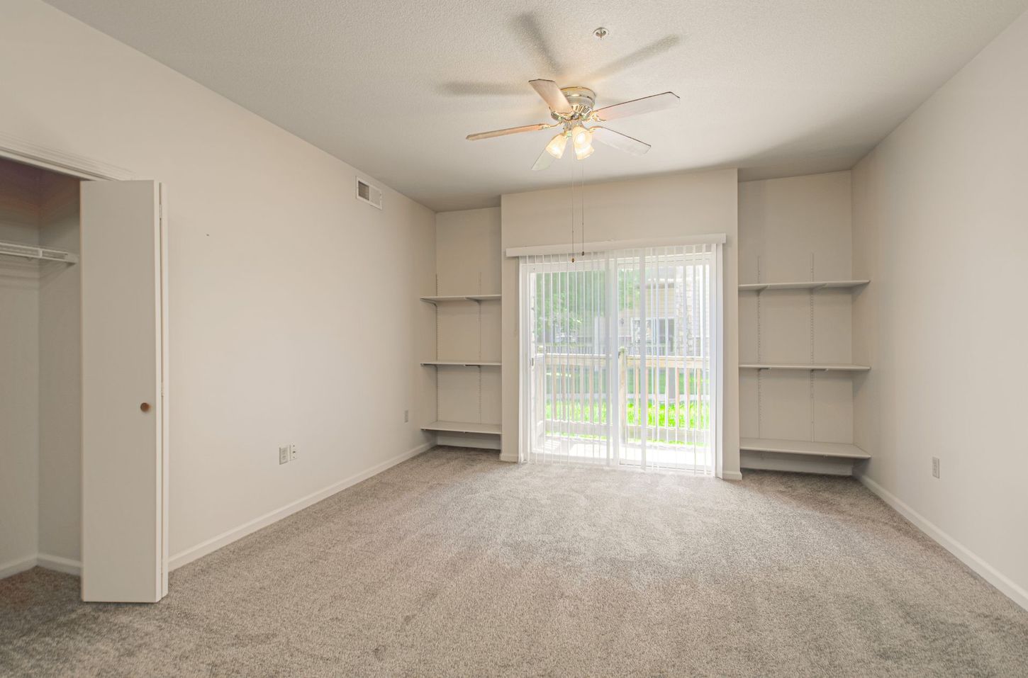 Empty bedroom with beige walls, carpet, and closet. Sliding glass door with shelves on either side and ceiling fan.