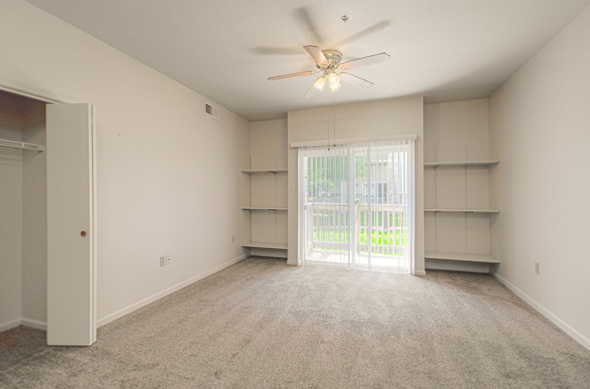 Empty bedroom with beige walls, carpet, and closet. Sliding glass door with shelves on either side and ceiling fan.