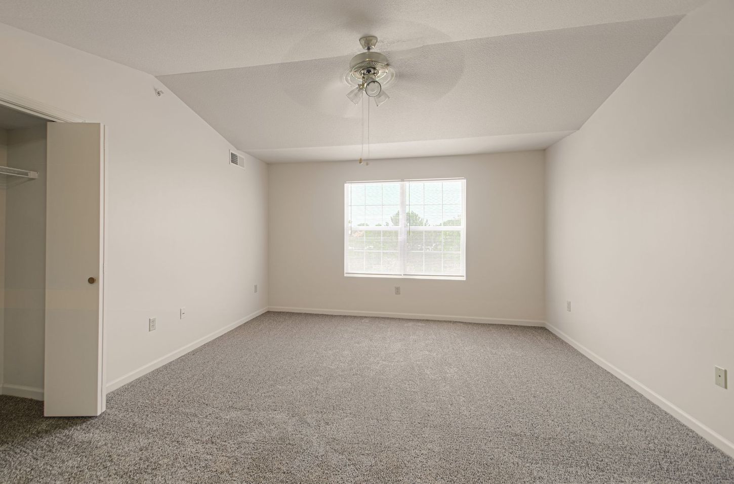 Empty bedroom with gray carpet, white walls, closet, and a window with a ceiling fan in the center.