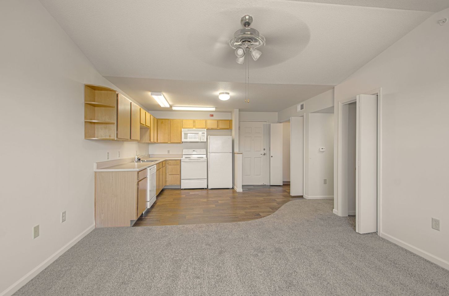 A small kitchen area with light-colored cabinets and appliances opens into a room with gray carpet and white walls.