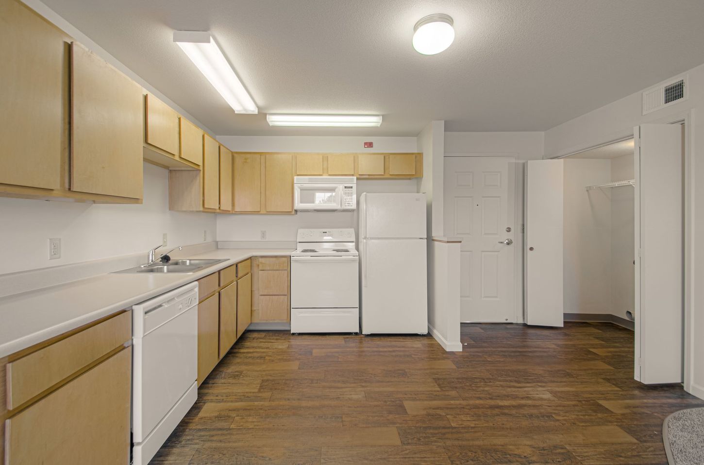 A kitchen with light brown cabinets and appliances. The floor is dark wood, and the walls are white.