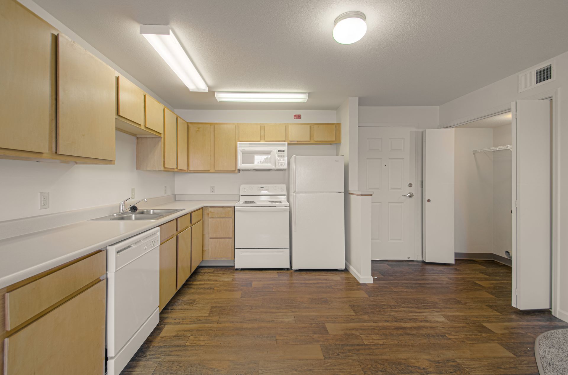 A kitchen with light brown cabinets and appliances. The floor is dark wood, and the walls are white.