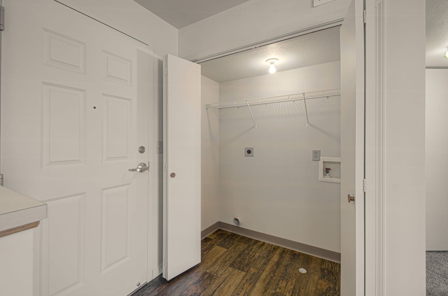 A utility room with a white door and open shelving. Dark wood-look flooring and a washing machine hookup are visible.