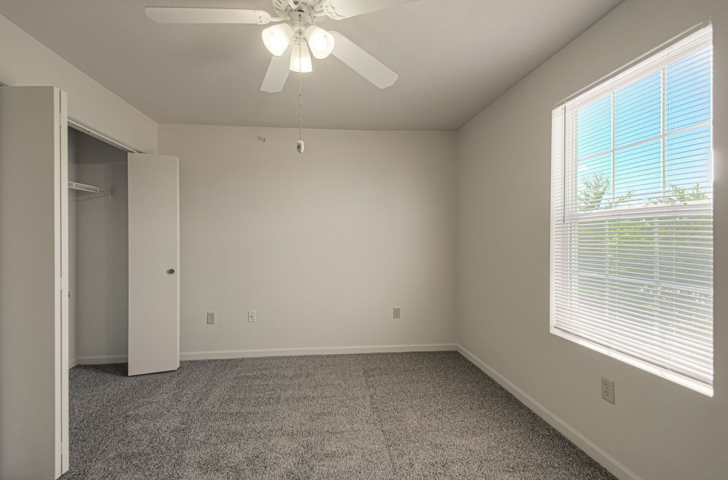 Empty bedroom with gray carpet, white walls, a closet, and a window with blinds. A ceiling fan is overhead.