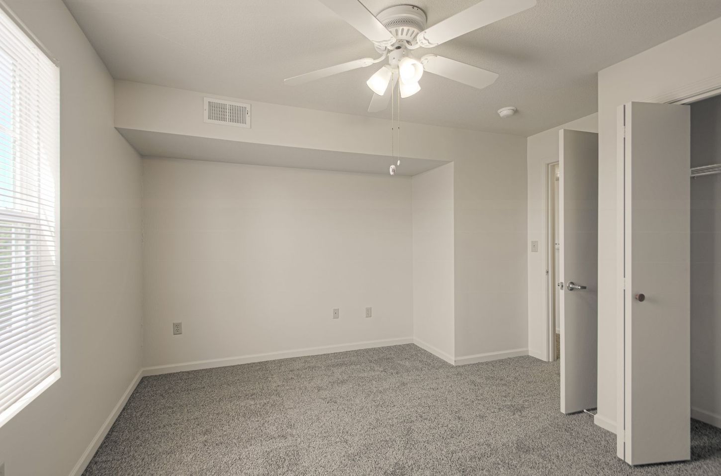 Empty bedroom with light-colored carpet, a ceiling fan, a window with blinds, and an open closet.