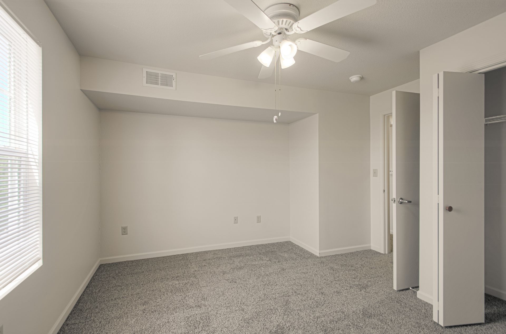 Empty bedroom with light-colored carpet, a ceiling fan, a window with blinds, and an open closet.