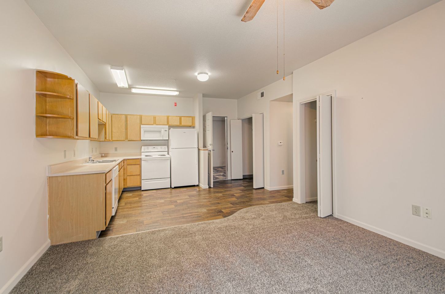 An empty apartment interior with a kitchen area, visible appliances, and doorways. The floor has varying flooring textures.