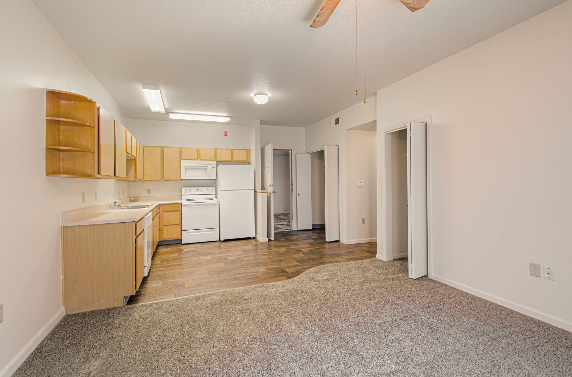 An empty apartment interior with a kitchen area, visible appliances, and doorways. The floor has varying flooring textures.