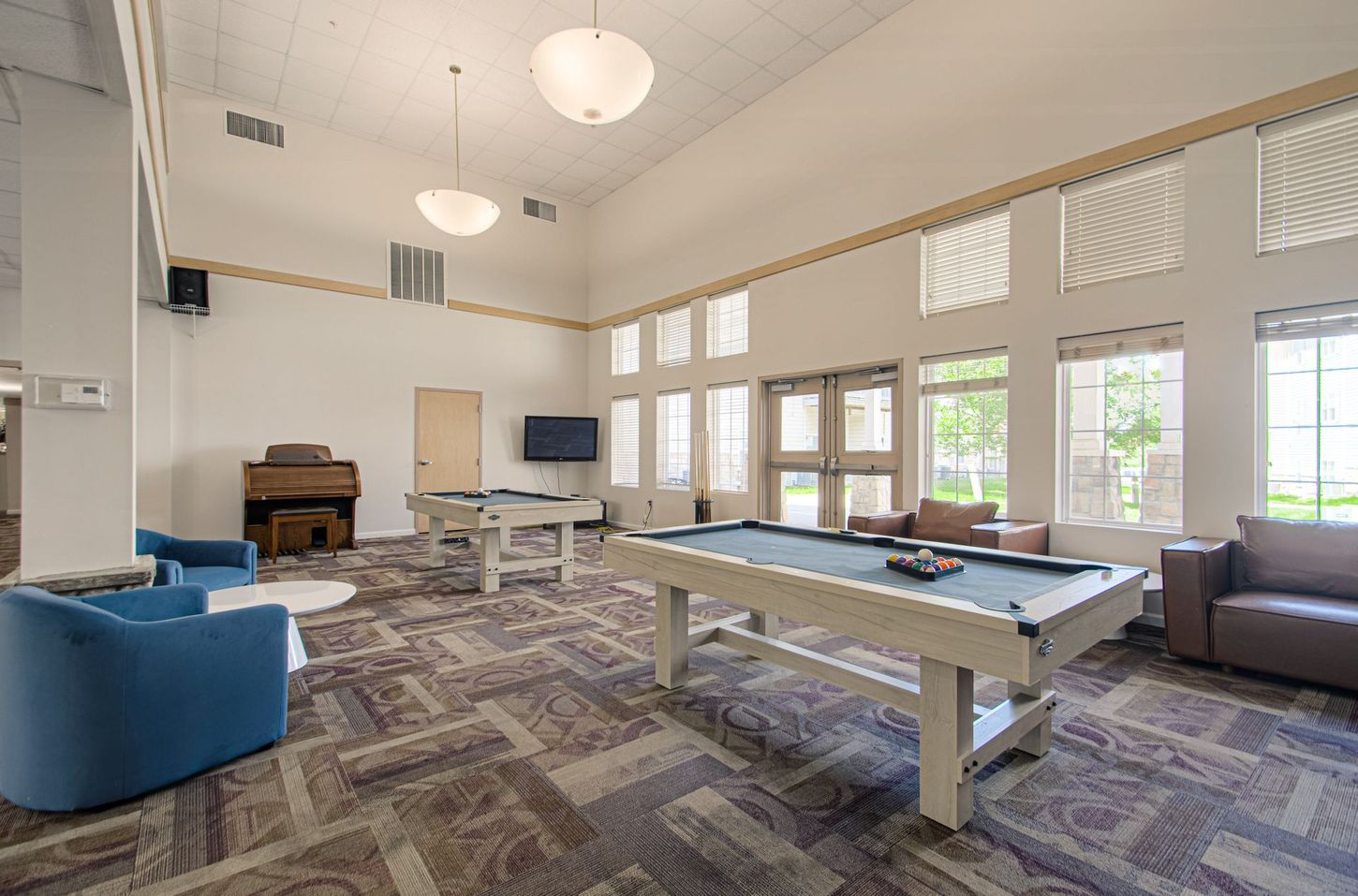 Spacious game room with two pool tables, lounge seating, and an upright piano, featuring neutral colors and natural light.