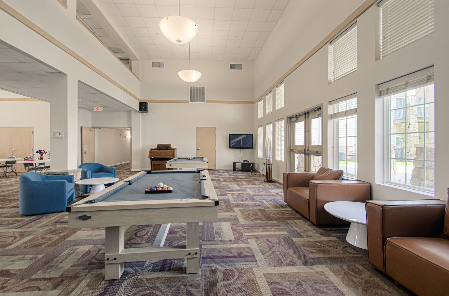 A community room with a pool table, seating areas, and large windows. The room is brightly lit with neutral tones and modern furnishings.