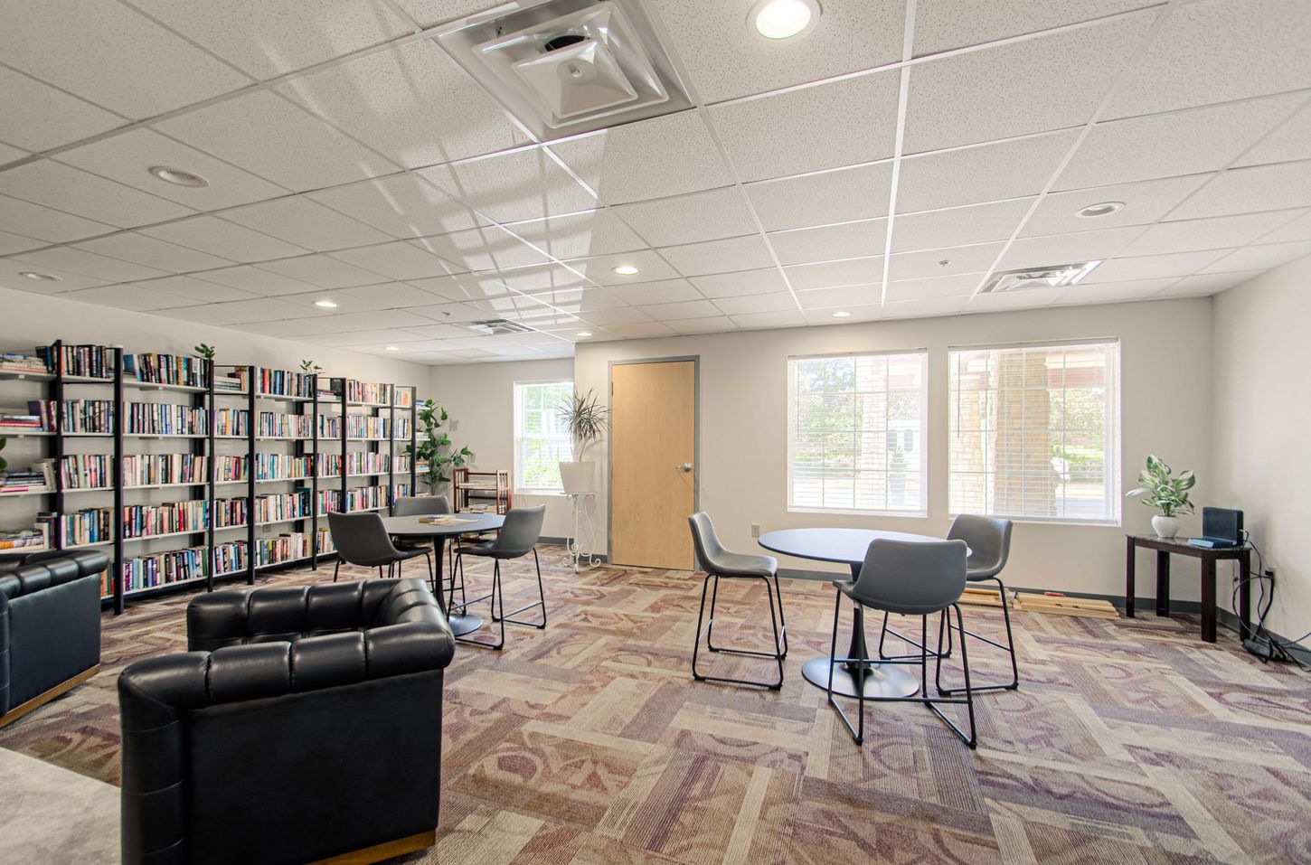 Cozy library area with bookshelves, tables, chairs, and a leather couch; featuring natural light from windows.
