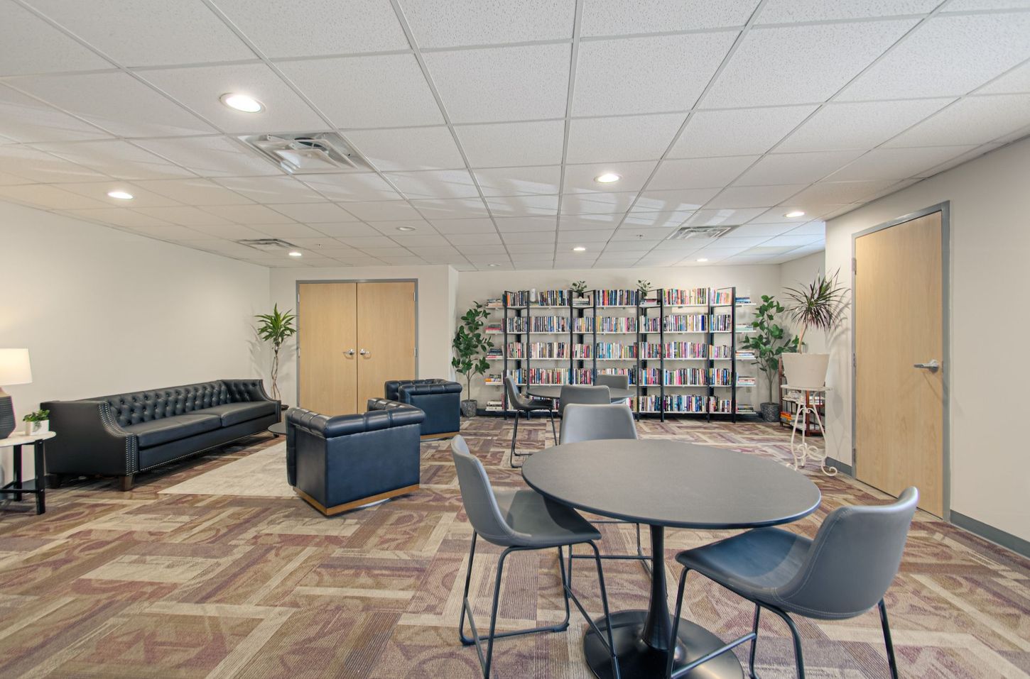 A modern lounge area with a round table and chairs, leather seating, and bookshelves. The space has a neutral color scheme with wood doors.
