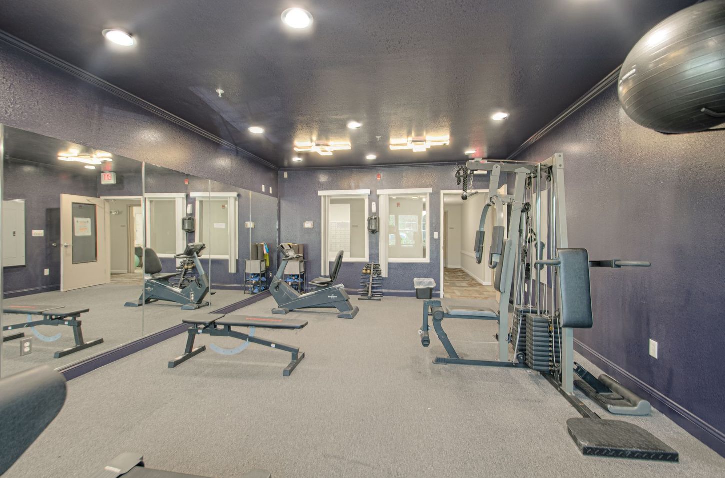 Gym interior with exercise equipment: weight machine, stationary bikes, weight bench, and mirrors. The walls are dark blue.