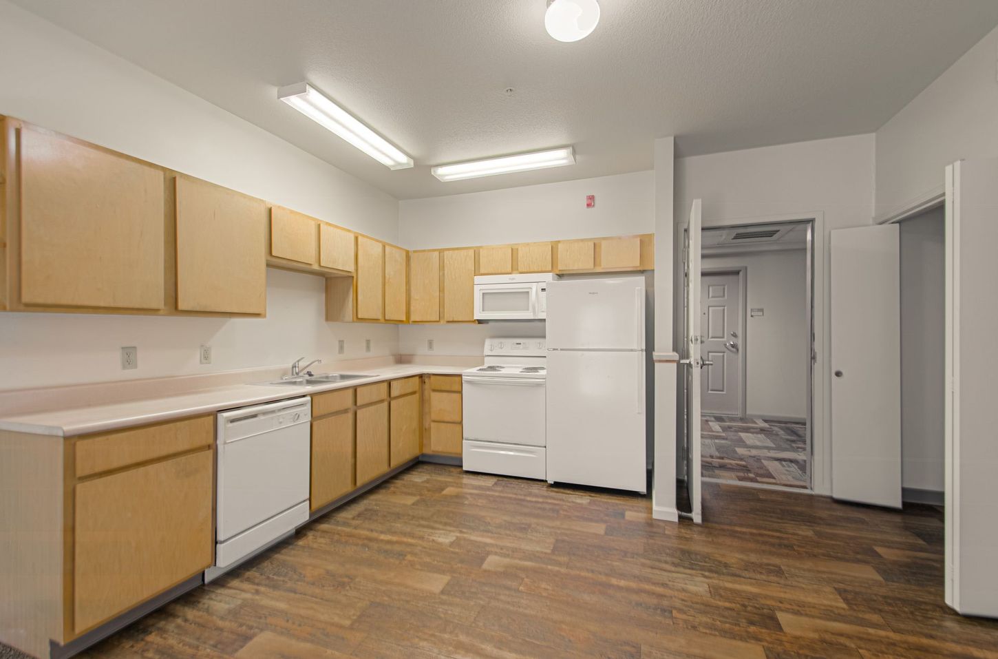 A kitchen with light-colored cabinets, white appliances, and brown flooring. An open doorway leads to a hallway.
