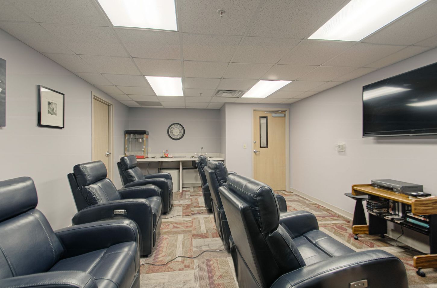 An empty media room with recliners, a large screen, and a snack station. The space has neutral tones and bright overhead lights.