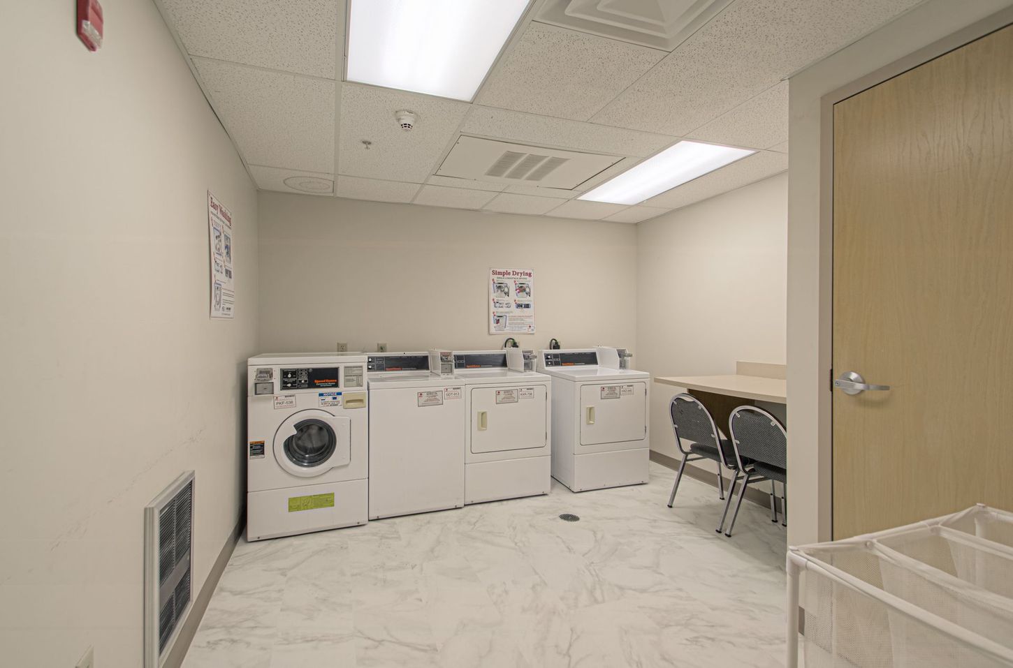 A laundry room with several white washing machines and dryers, a table with chairs, and a light-colored door.