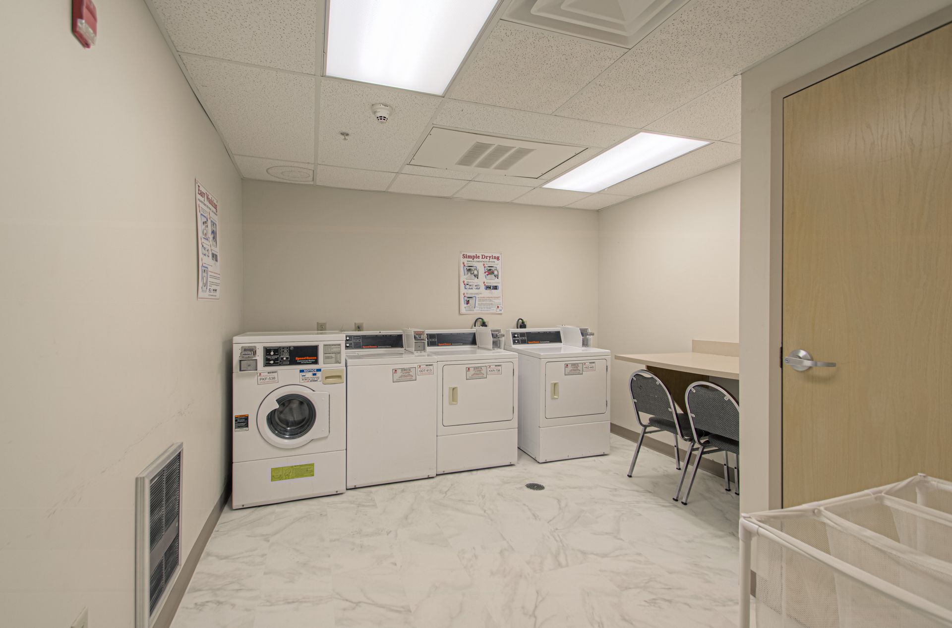 A laundry room with several white washing machines and dryers, a table with chairs, and a light-colored door.