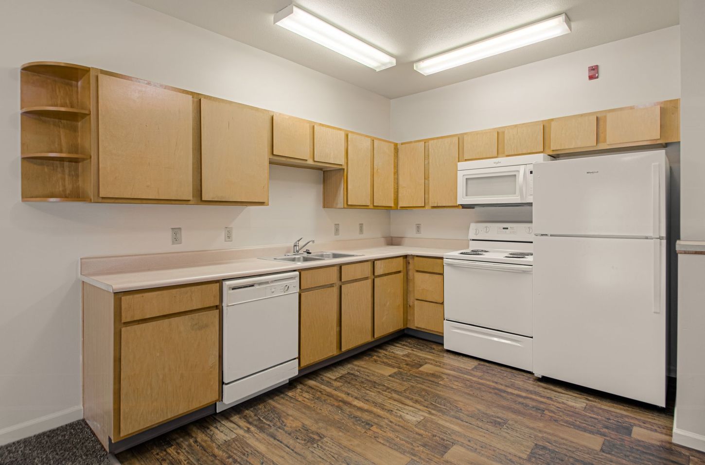 A small kitchen with light wood cabinets, white appliances, and a beige countertop.  The floor is wood-look laminate.