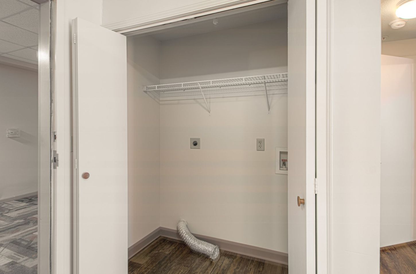 Laundry closet with white doors open, showing a shelf, outlets, and a vent hose on the floor.