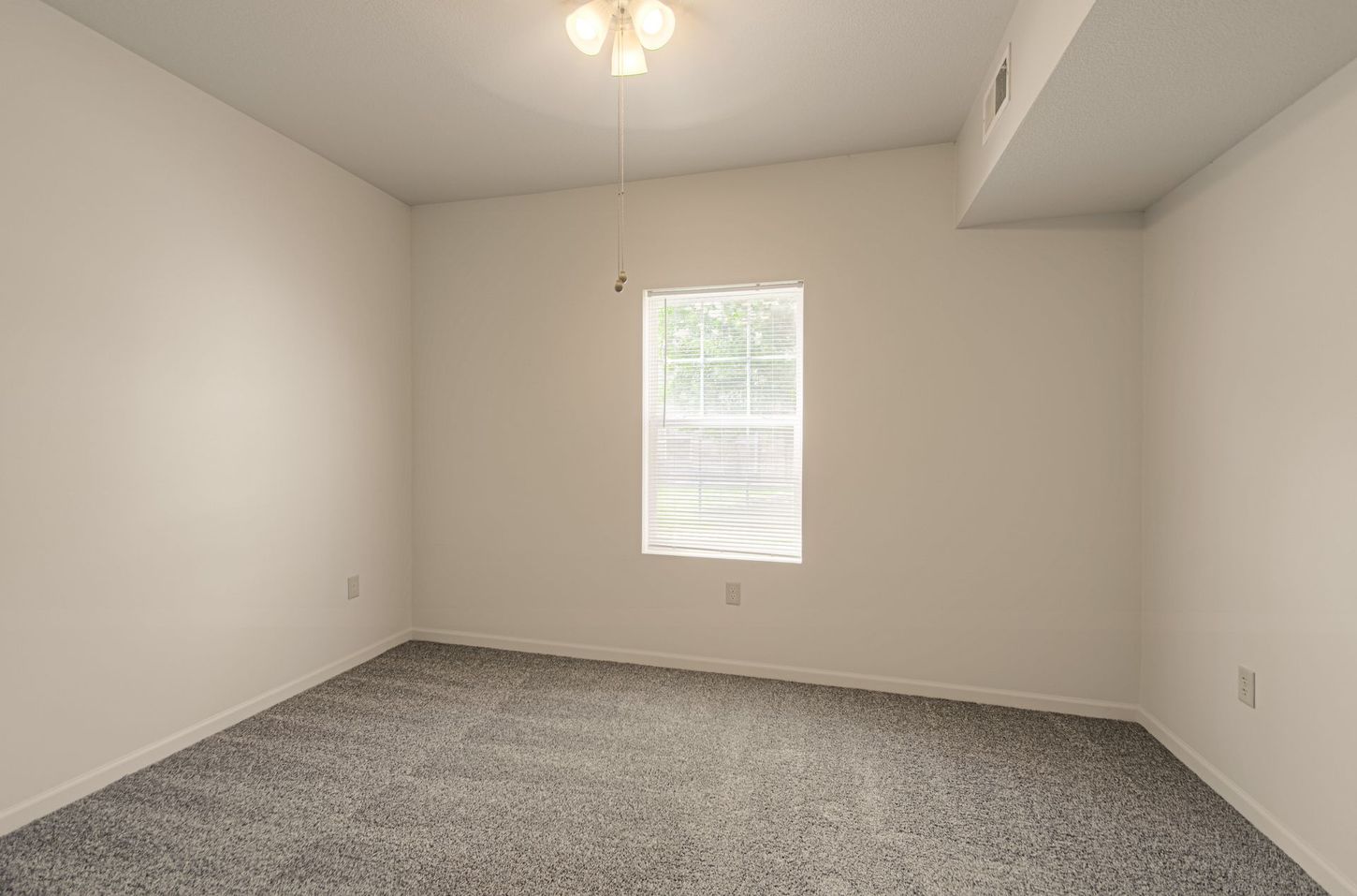 Empty room with gray carpet, white walls, and a window with blinds. A ceiling fan and overhead light fixture are visible.