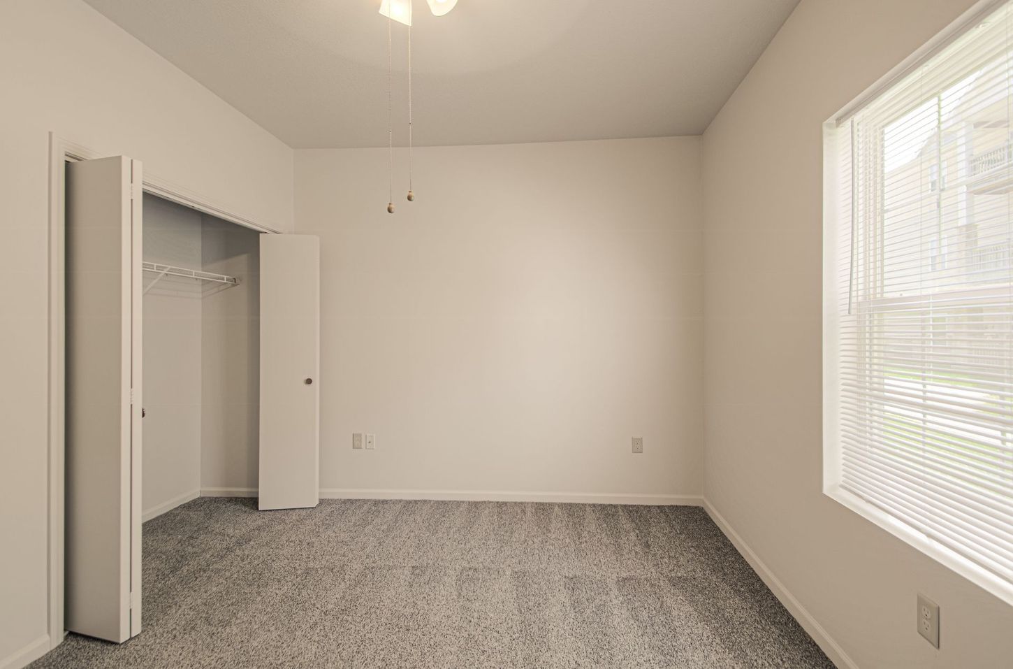 Empty bedroom with gray carpet, closet, and window with blinds. White walls and a ceiling fan.