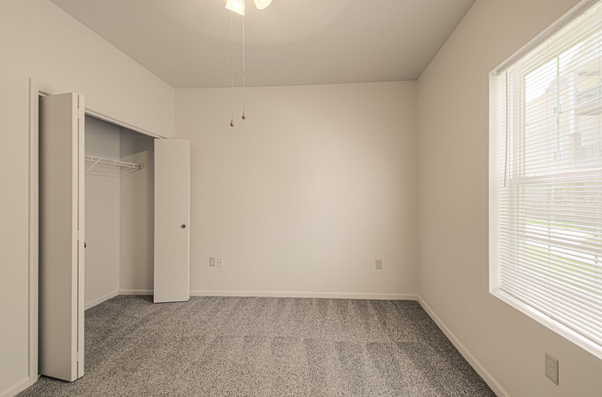 Empty bedroom with gray carpet, closet, and window with blinds. White walls and a ceiling fan.