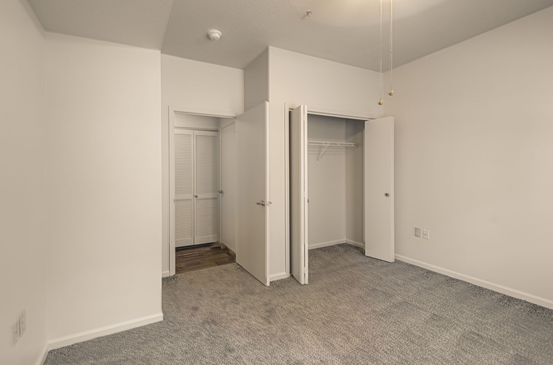 Empty bedroom with grey carpet, white walls, and two closets. One closet door is open.
