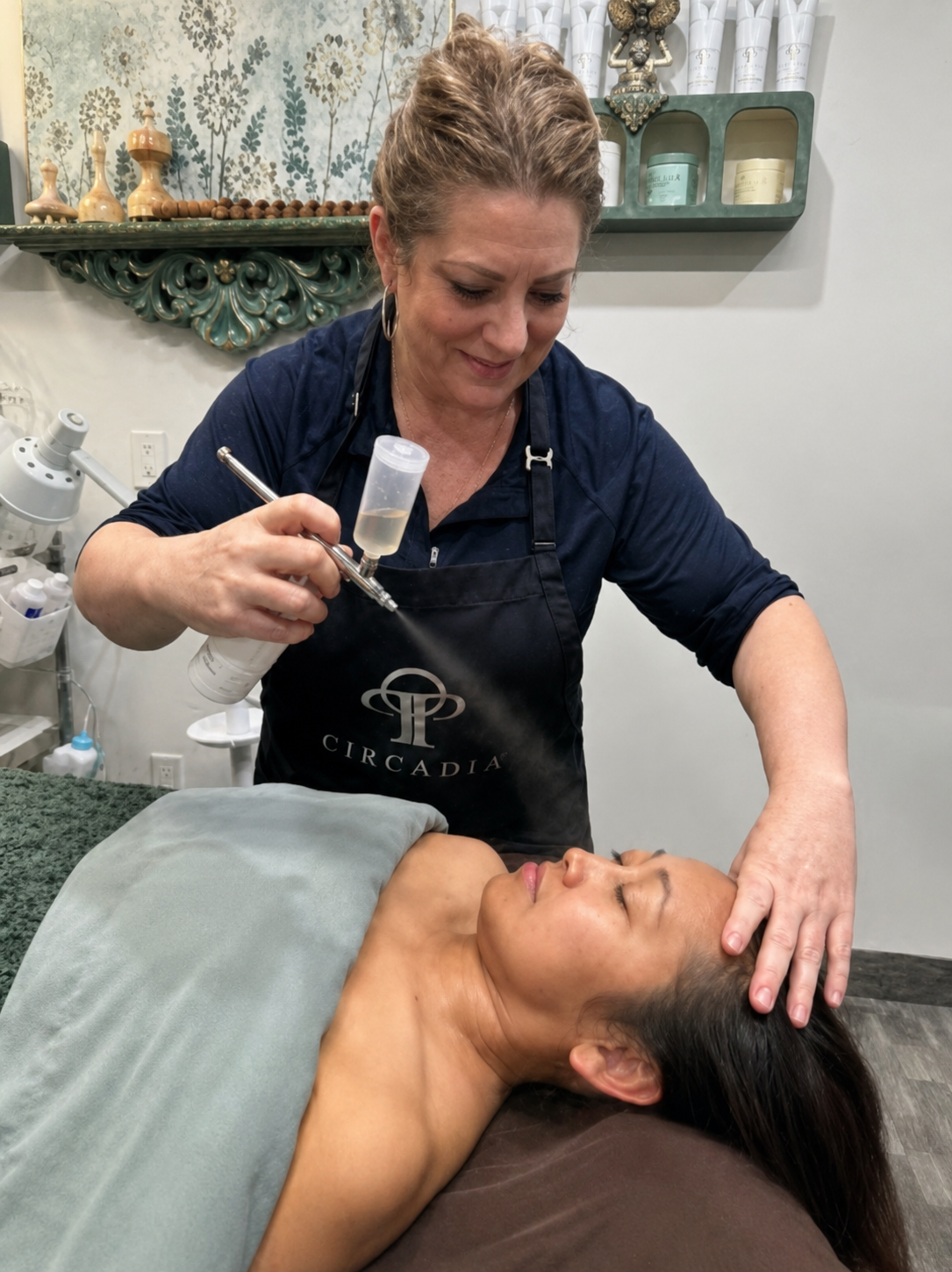 A person in a black uniform uses an airbrush tool on a client receiving a facial treatment in a salon setting.
