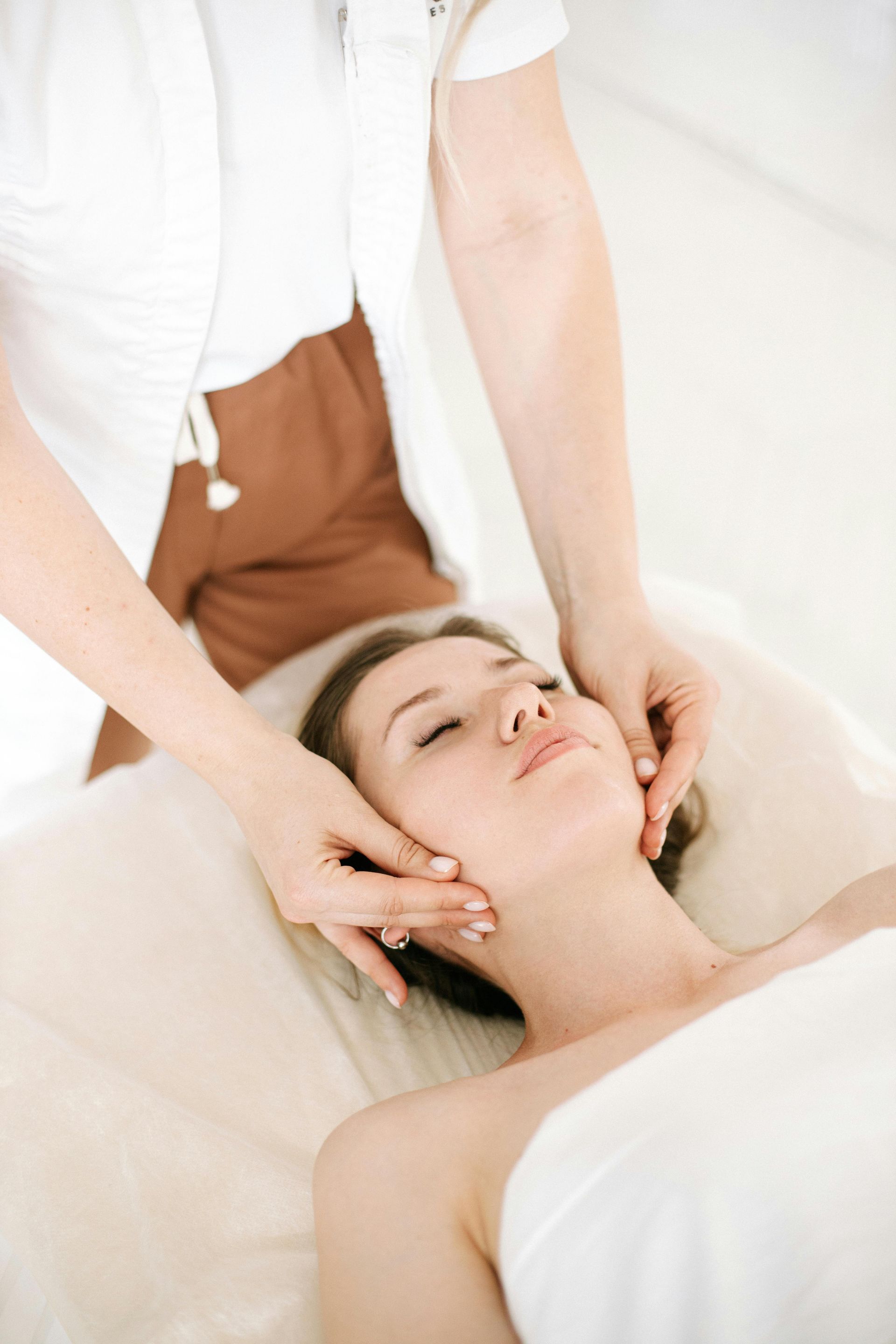 Woman receiving facial massage in a spa. Hands on cheeks, lying down, white towel, light setting.