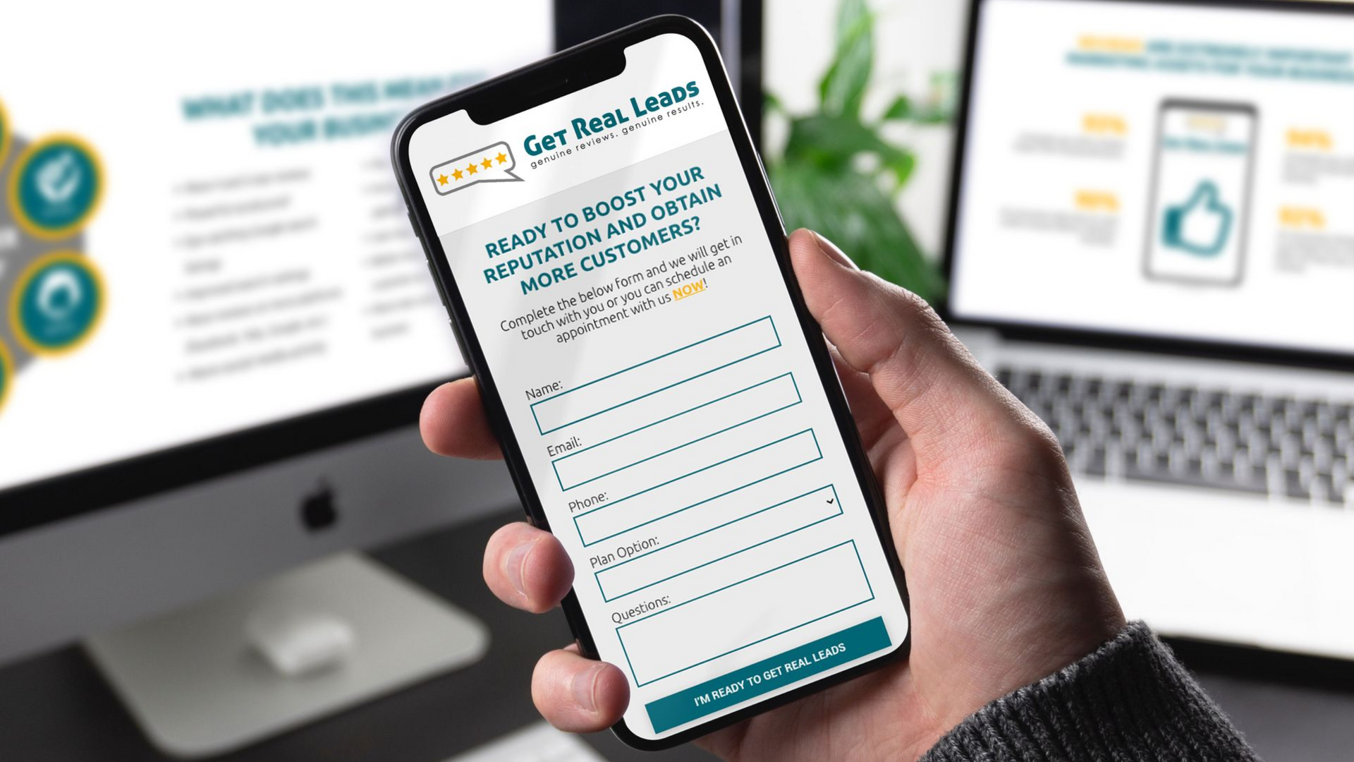 Person holding a phone with a web form open, with a computer screen and laptop in the background.