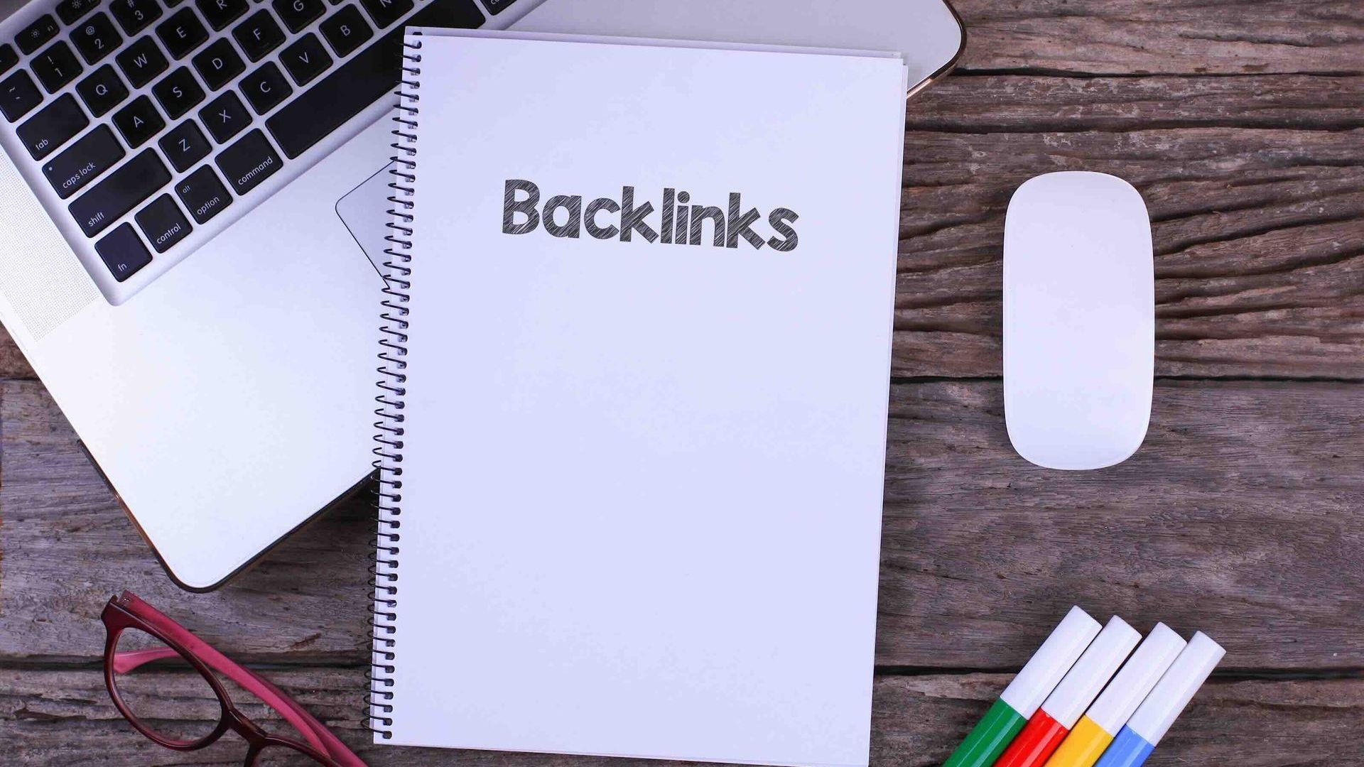 A notebook with the word backlinks written on it is sitting on a wooden table next to a laptop.