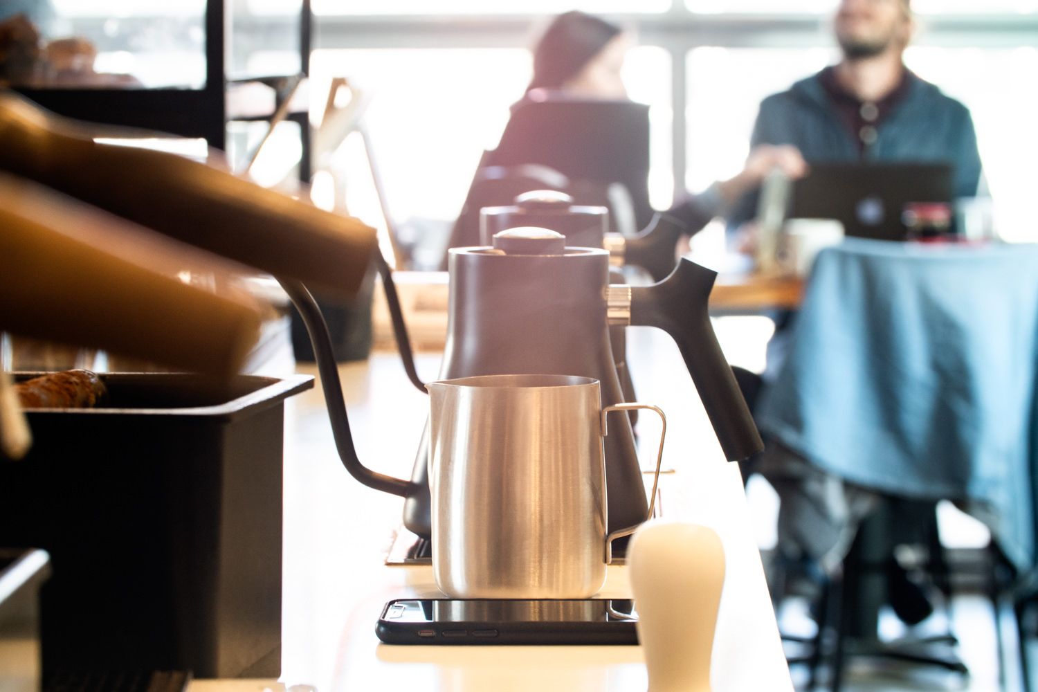 Coffee brewing station in a cafe, stainless steel kettle, barista in the background.