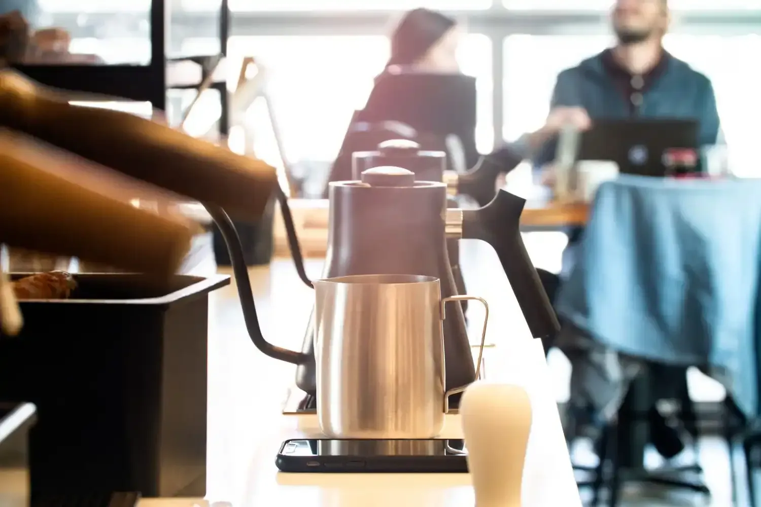 Coffee brewing station in a cafe, stainless steel kettle, barista in the background.