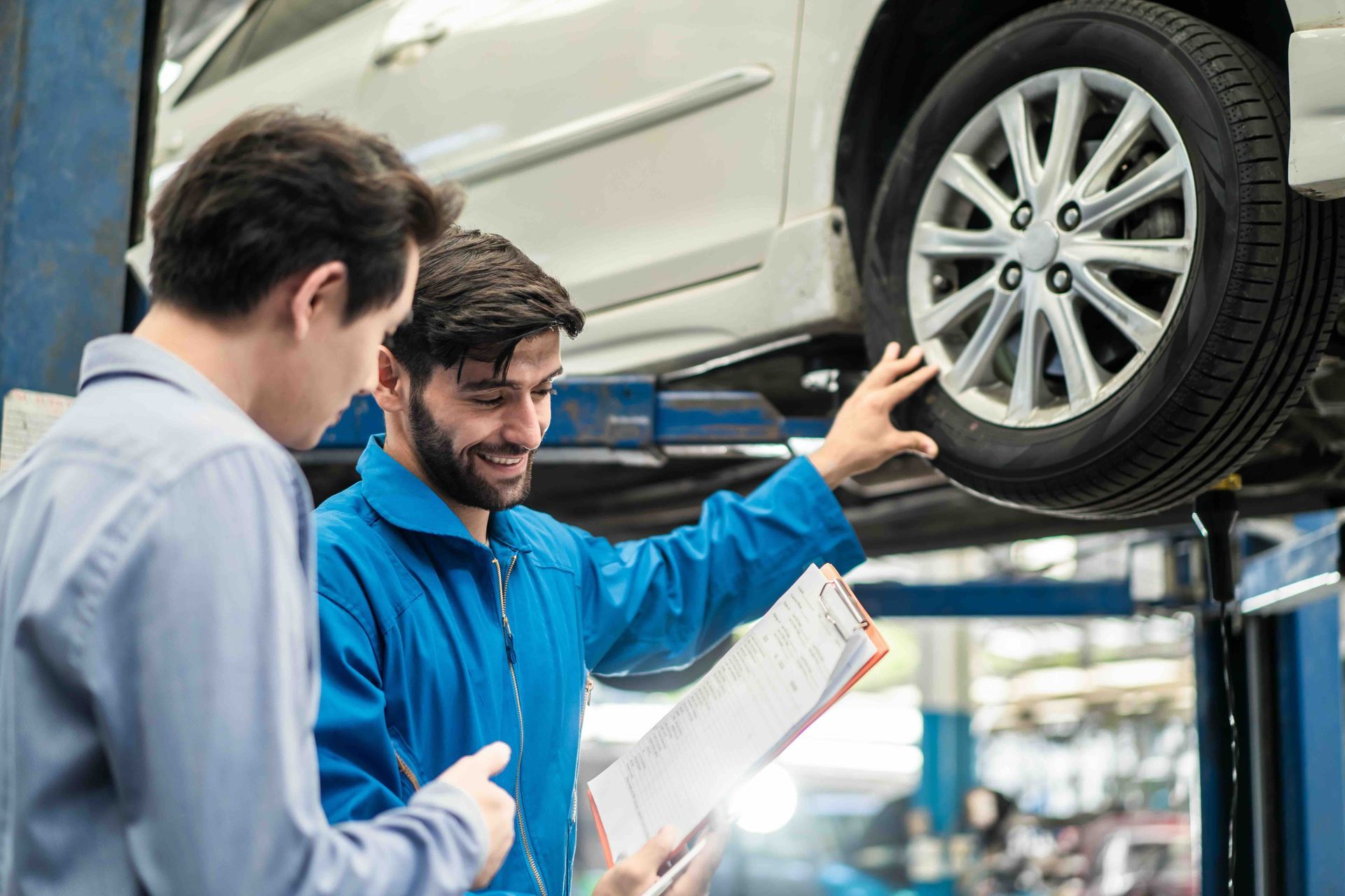A mechanic is showing a car's wheel to a customer with a clipboard in hand, inside a garage.