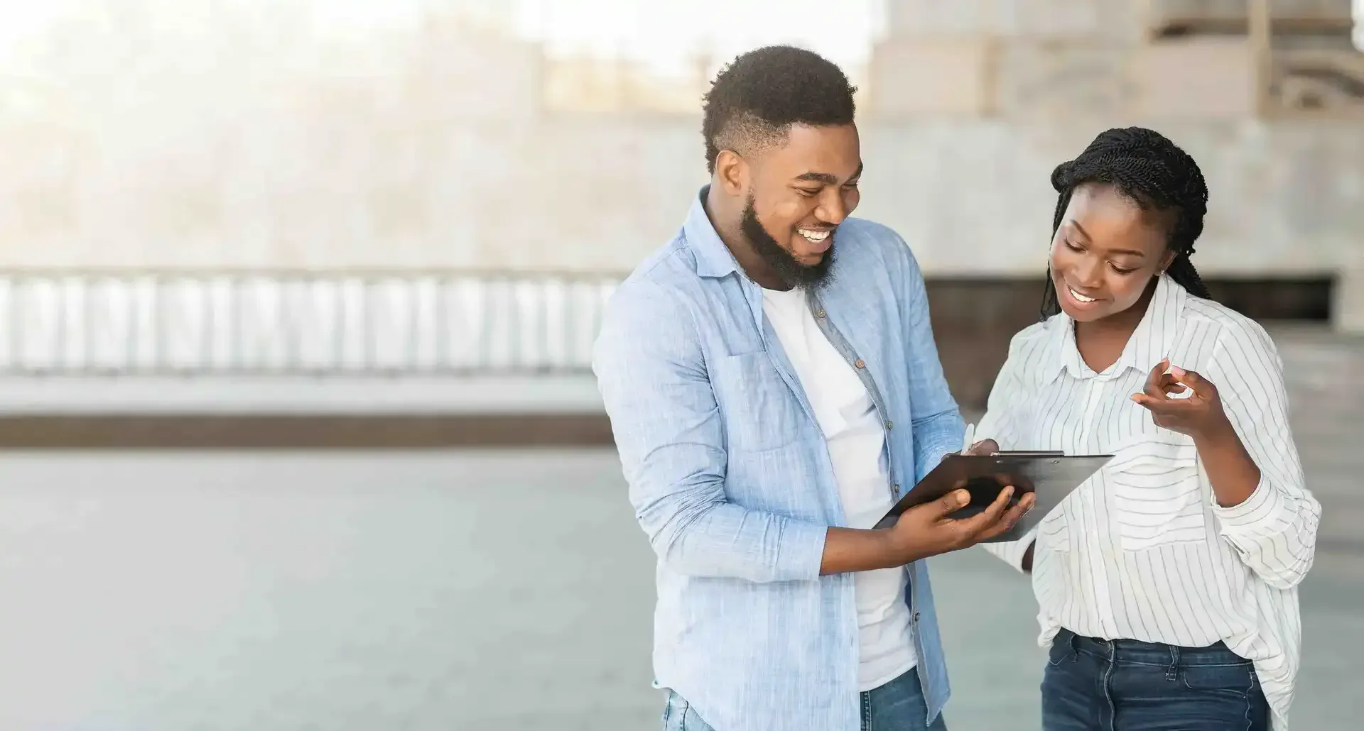 A black male and female are smiling and looking at a tablet while outsde.