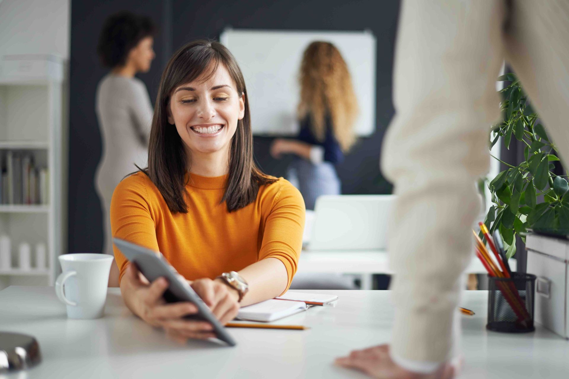 A woman is sitting at a desk is pointing and showing someone else her tablet computer screen.