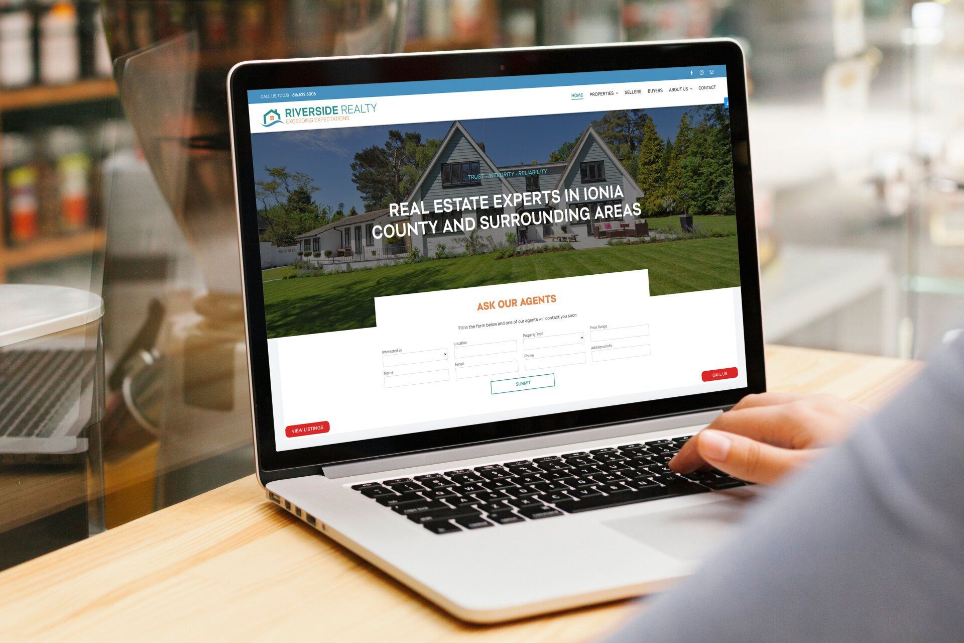 A person is searching Riverside Realty's website on a laptop computer on a wooden table.