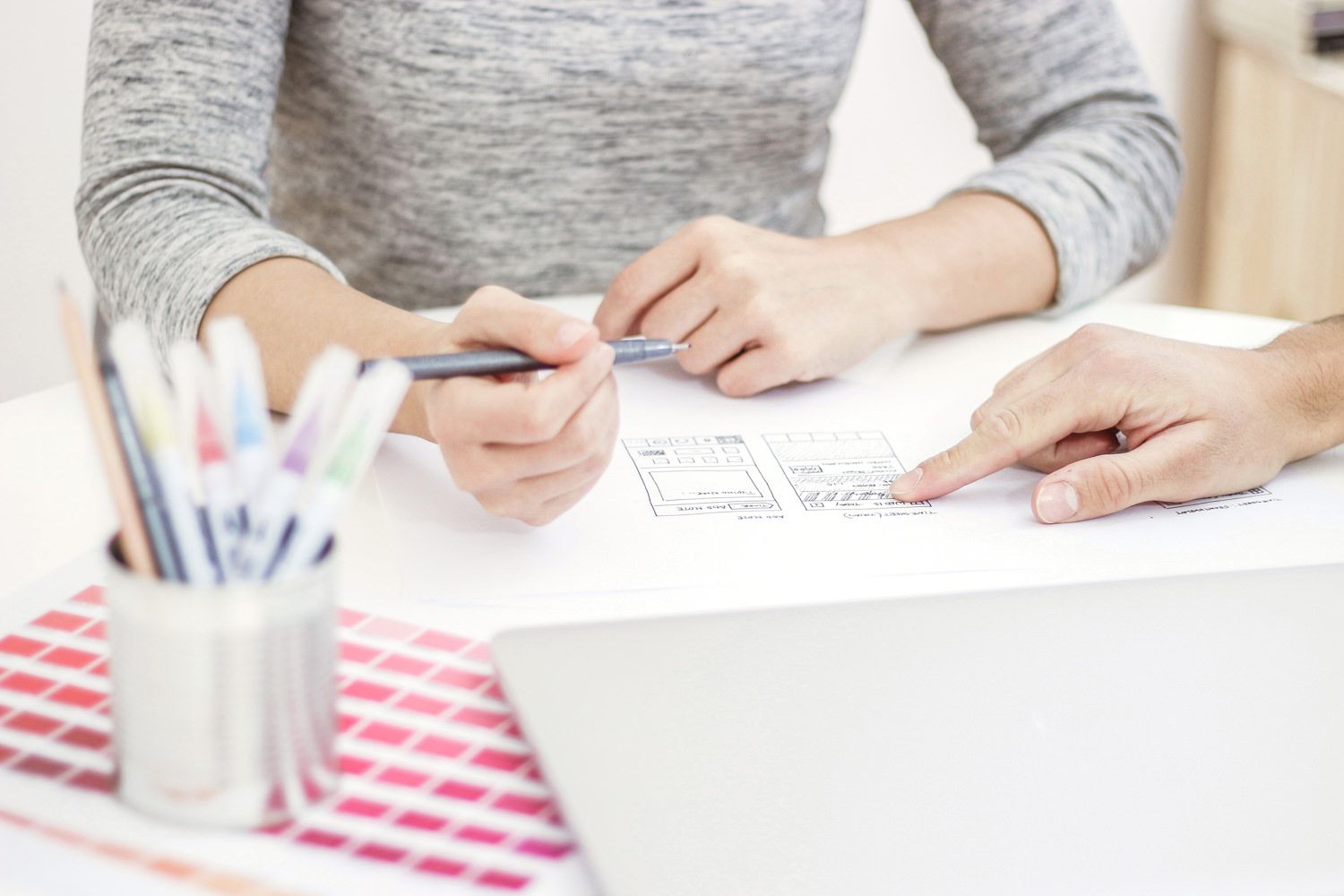 Two people collaborate on a design layout, pointing and drawing, with art supplies and a laptop on a table.